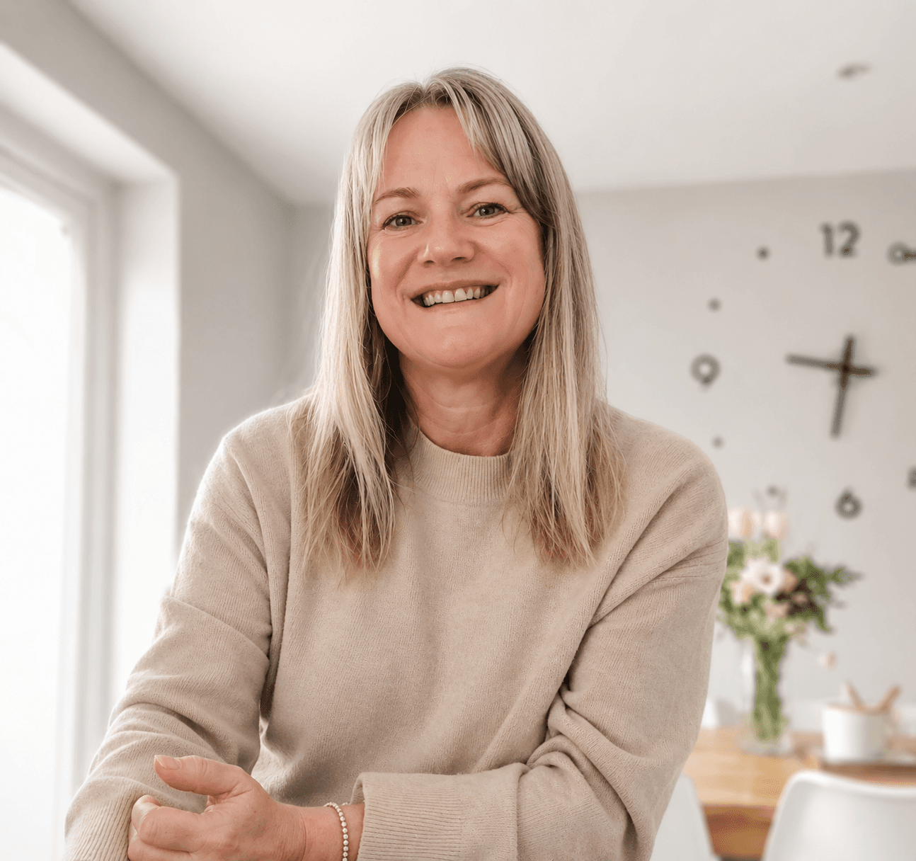 Smiling middle-aged woman with blonde hair wearing a beige sweater in a bright home.